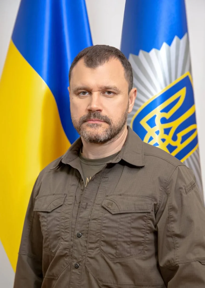 Чоловік з бородою у коричневій сорочці на тлі українських прапорів